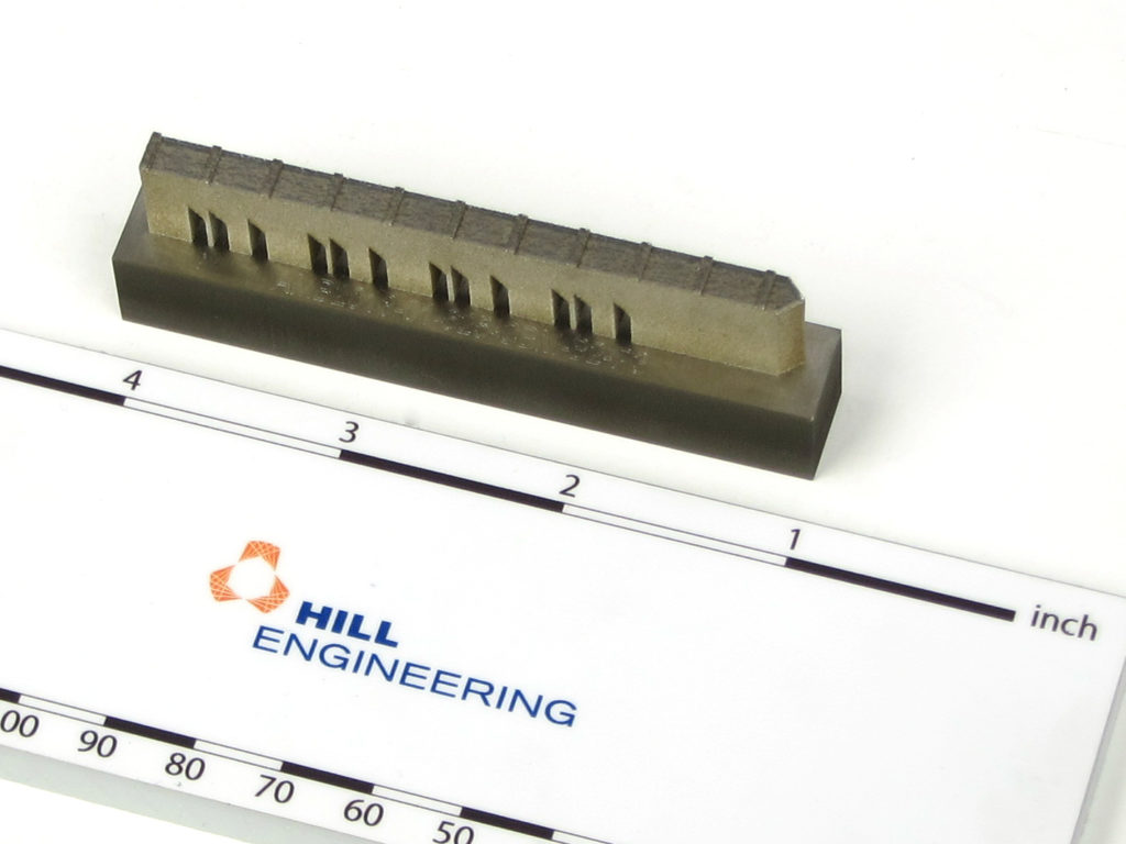 Photograph of an Additive Manufacturing Benchmark test specimen next to a ruler containing a Hill Engineering logo