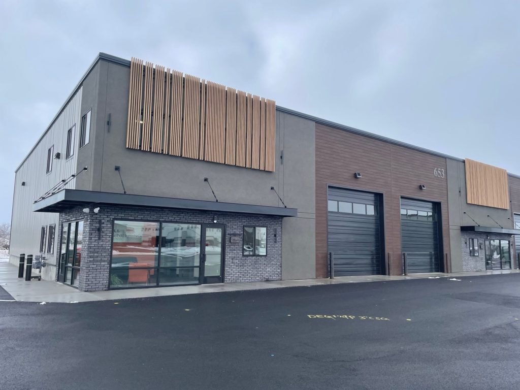 Gray building with brick and wood exterior accents and two black roll up warehouse doors against an overcast sky.