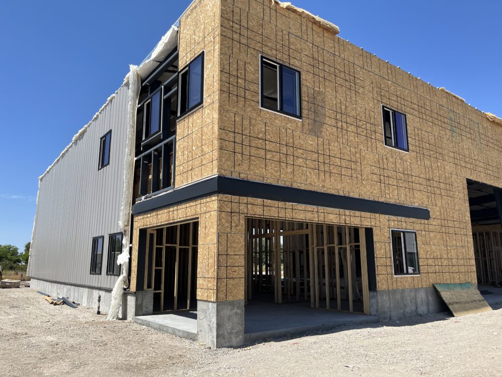 Gray building with in-progress construction on the front against a blue sky. Wood framing can be seen in the interior and particle board sheets are on the exterior.