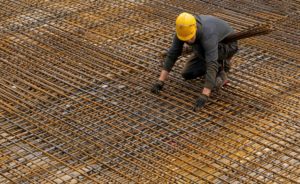 A construction worker wearing a yellow hard hat crouched over a grid of metal rebar.