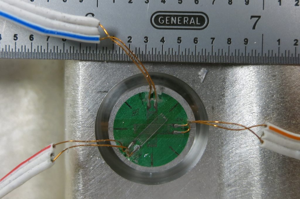 A ring cut into an aluminum surface circumscribing a green strain gage with thin wires attached.