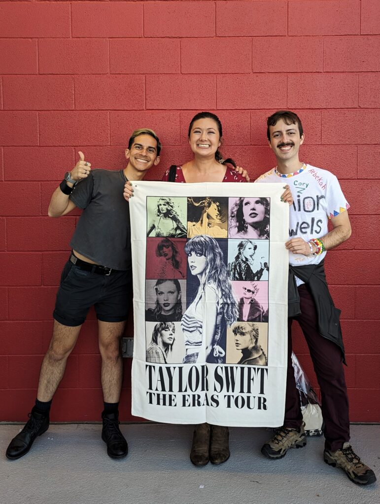Robby, Camille, and Ryan hold a Taylor Swift Eras Tour tapestry while standing in front of a red brick wall.