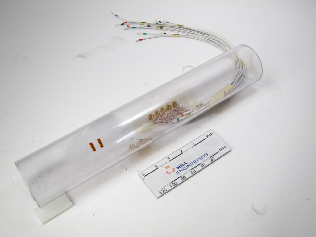 A clear open-ended cylinder with strain gages on the inner diameter. White wires are attached to the strain gages and come out of one end.