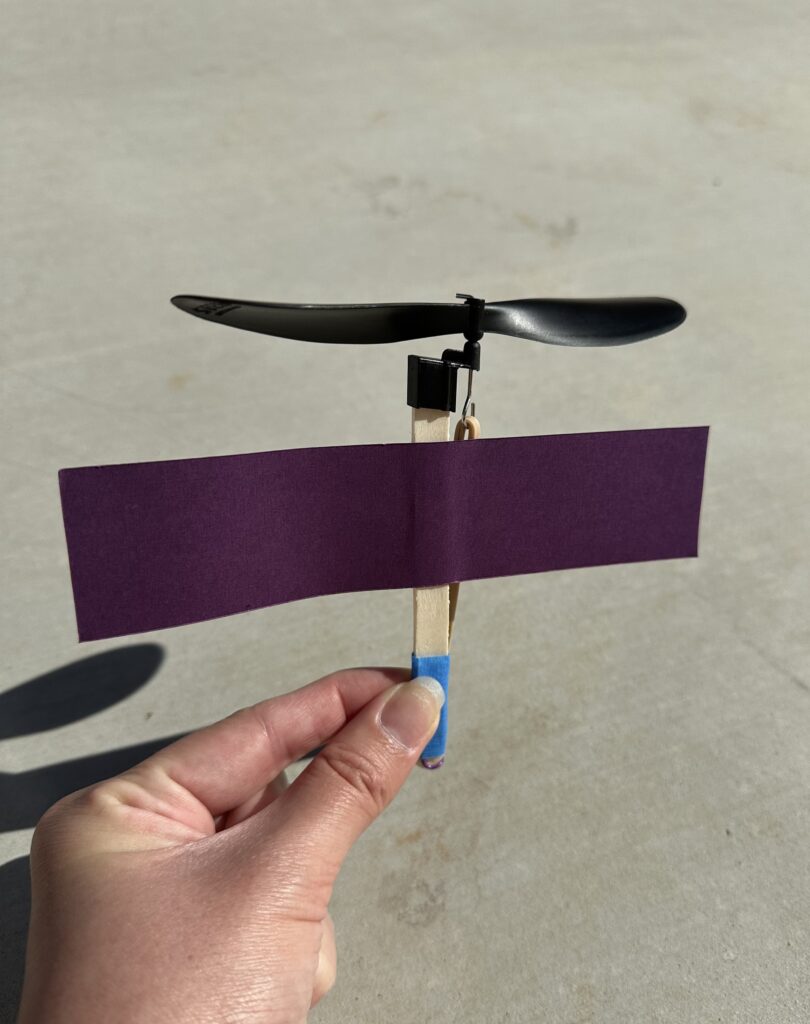 A hand holding a helicopter made from a popsicle stick, rubberbands, tape, and a rectangular purple piece of paper for the body.