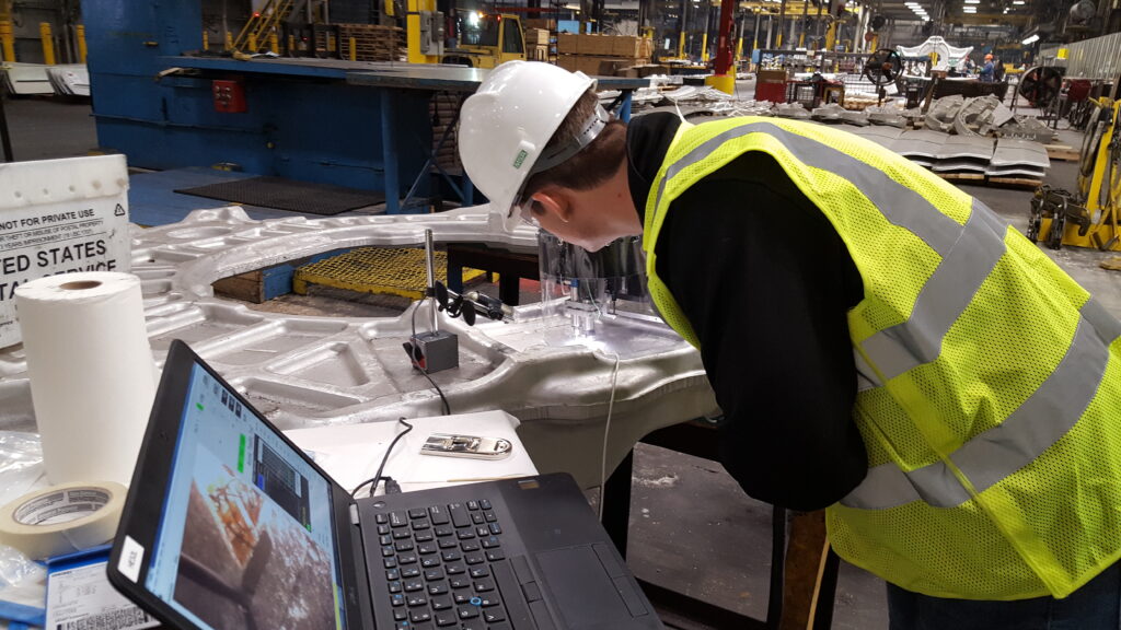 A worker in a yellow safety vest and white hard hat inspecting a metal forging with measurement equipment in a factory setting. A laptop is in the foreground showing camera footage of the measurement site.