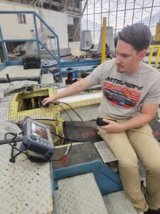 A person sitting on top of an A-10 aircraft in a hangar performing an inspection with a handheld tool while looking at a monitor