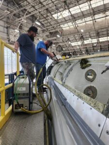 Two people standing on scaffolding next to an A-10 aircraft in a hangar performing maintenance with a hand held tool