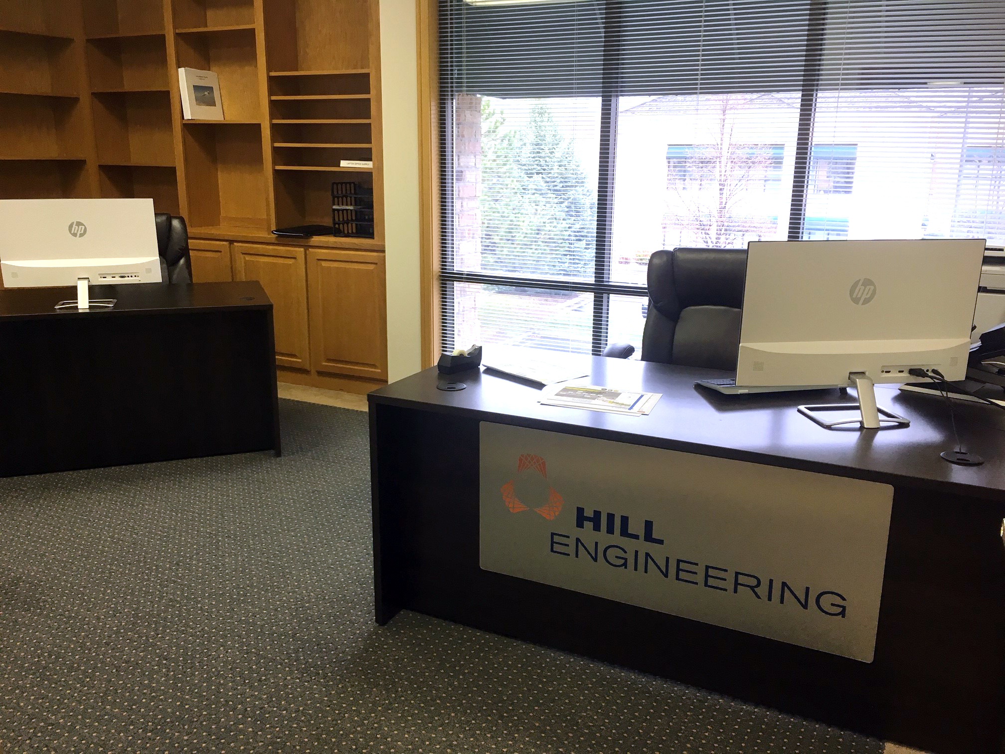 The interior of an office with a desk that reads "Hill Engineering" on the front and several bookshelves in the background.