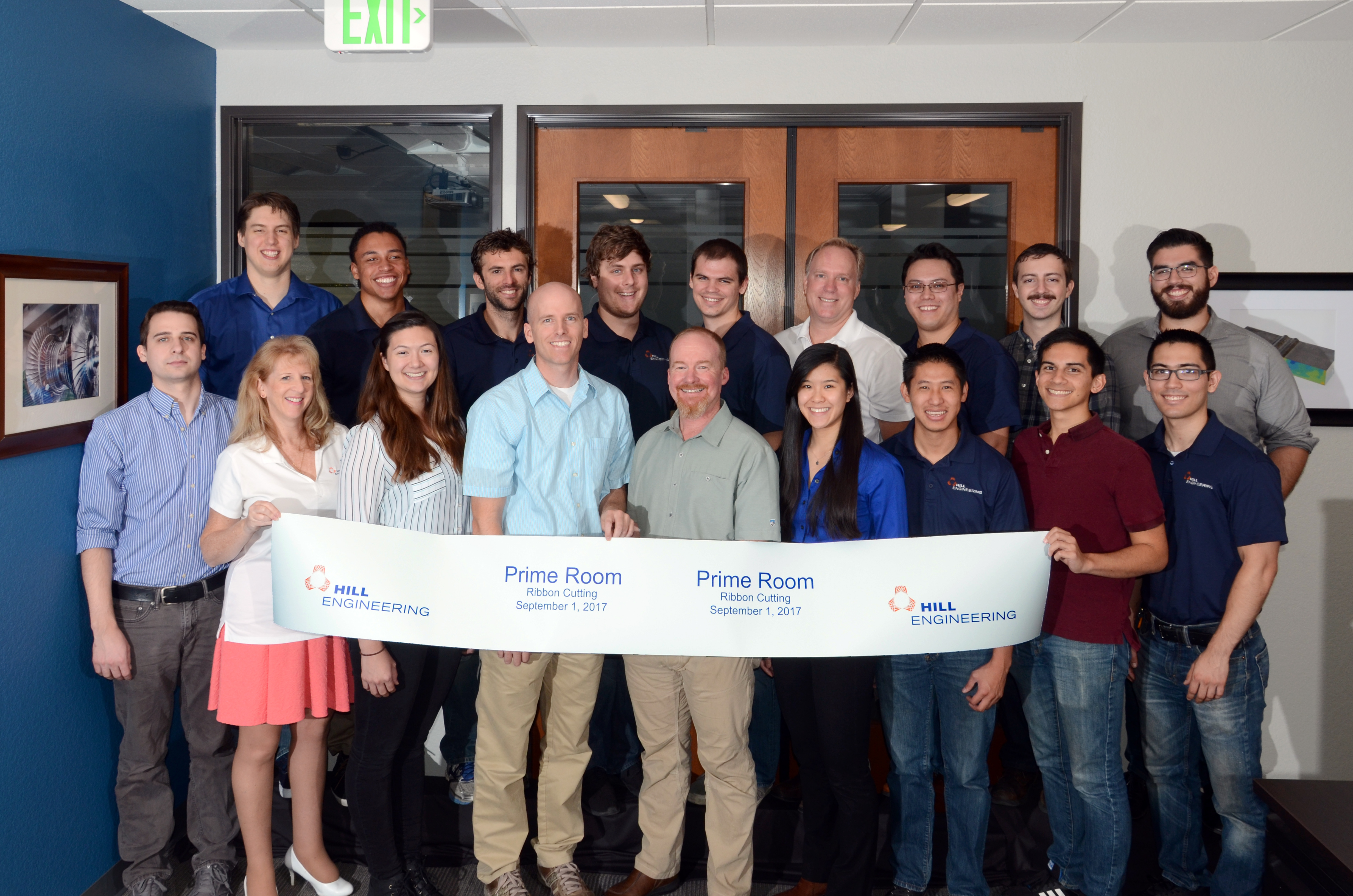A group of 18 adults standing in two rows holding up a sign commemorating the "Prime Room."