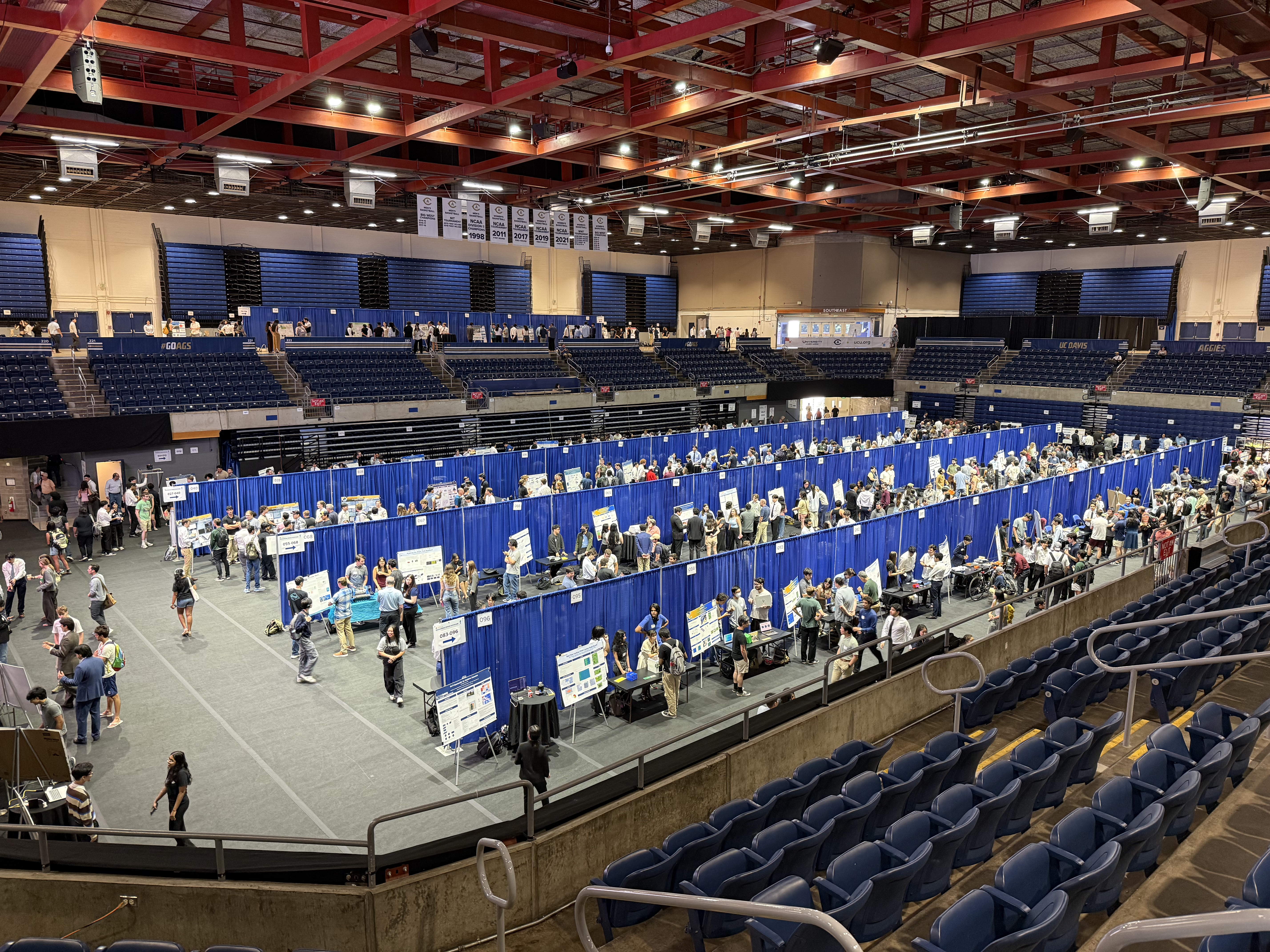A large auditorium with the floor level separated into rows by hanging blue curtains. Within the rows are presentations featuring a table display and poster hosted by small groups of university students for their senior design project. Evaluators roam through the presentations.